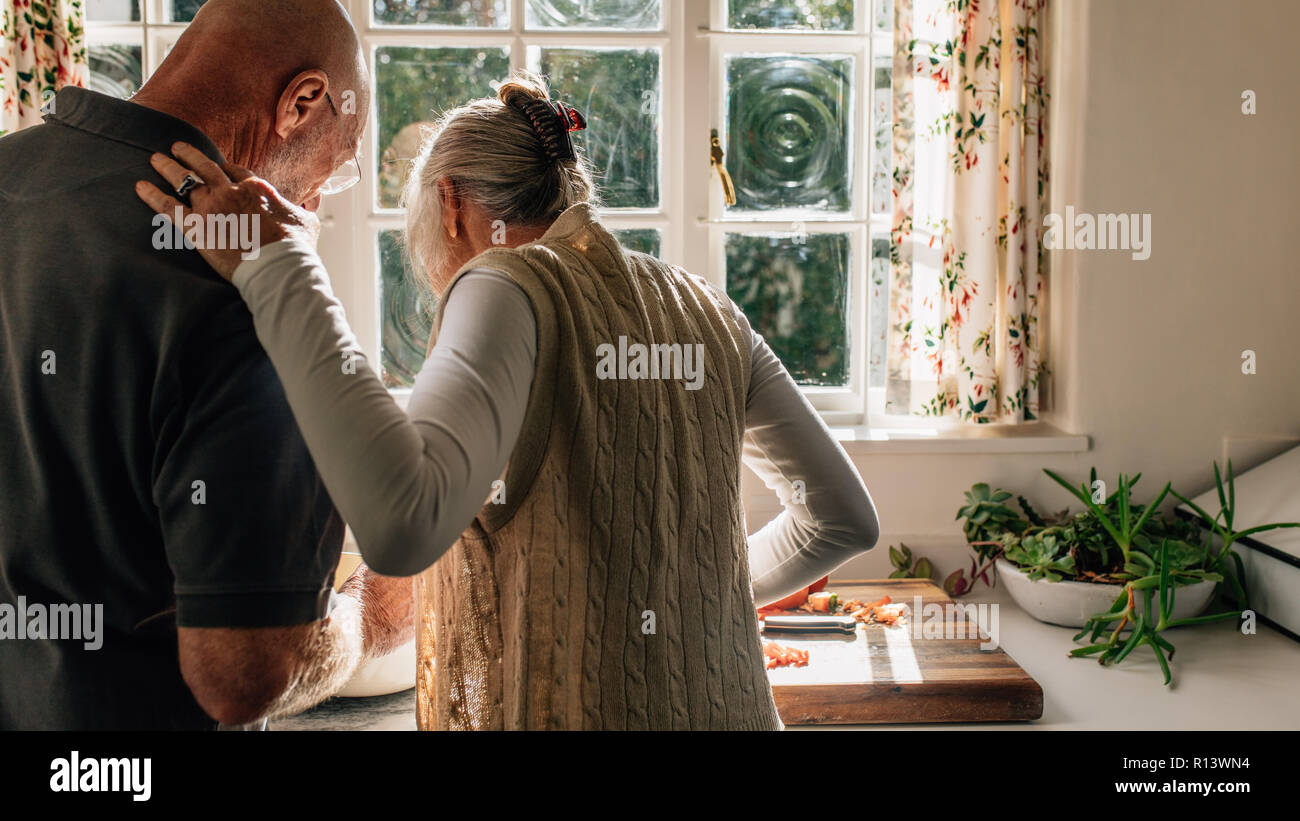 Vista posteriore di una coppia senior in piedi insieme nella cucina di cottura degli alimenti. Donna anziana guardando suo marito cucinare il cibo in piedi in cucina. Foto Stock