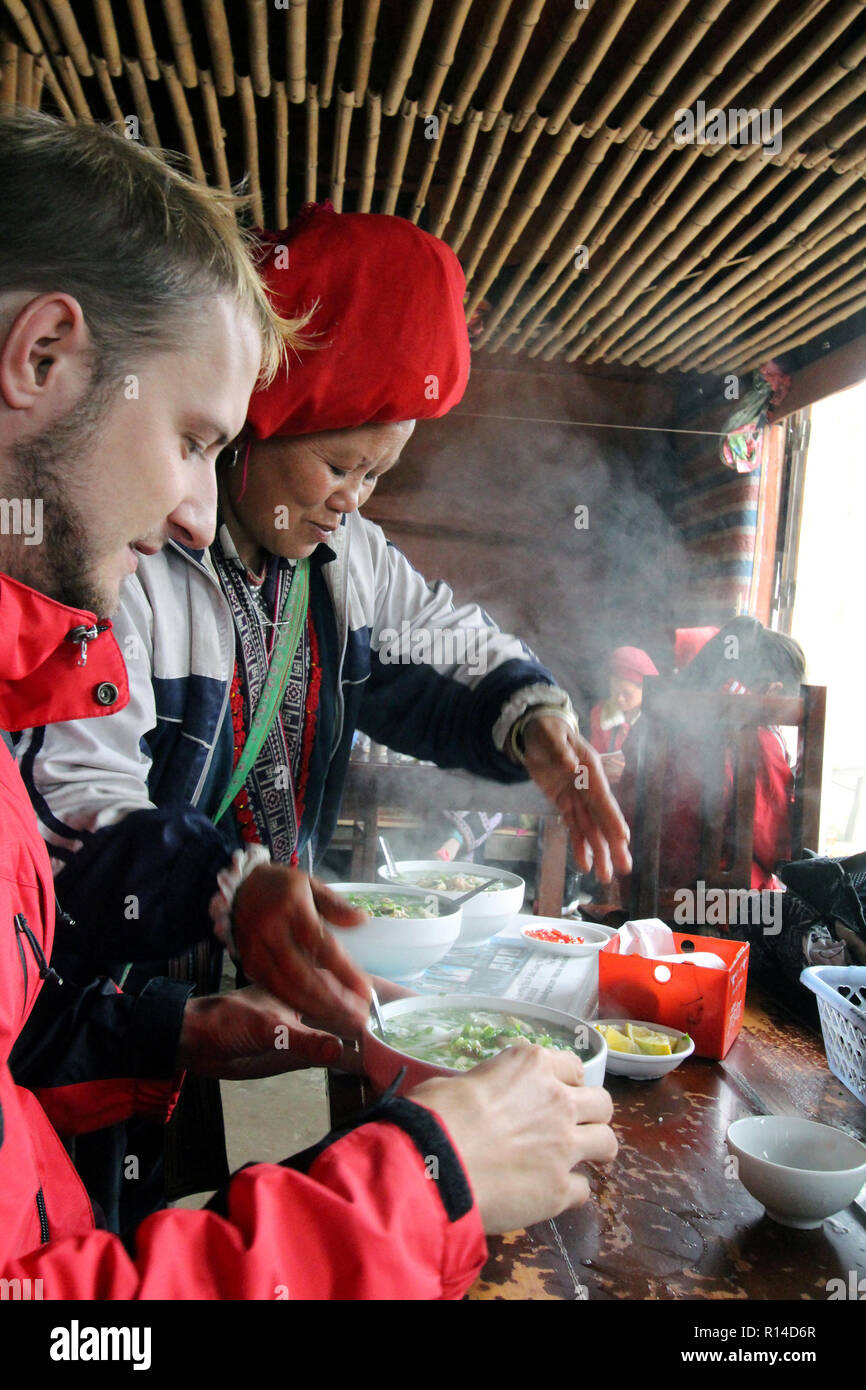 Red dzao donna serve pho ga (zuppa di noodle al pollo) per un turista tedesco al suo villaggio eatery vicino a SAPA, Vietnam Foto Stock