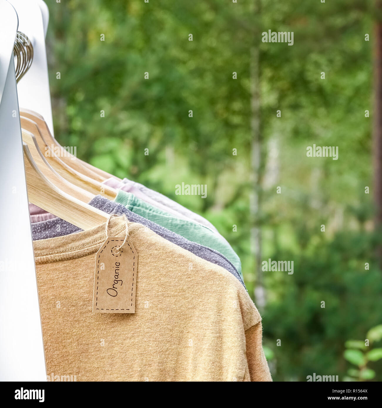 Abbigliamento organico. Colore naturale t-shirts appesi i portabiti in legno in una fila. Eco tag tessili. Verde bosco, Natura in background. Foto Stock