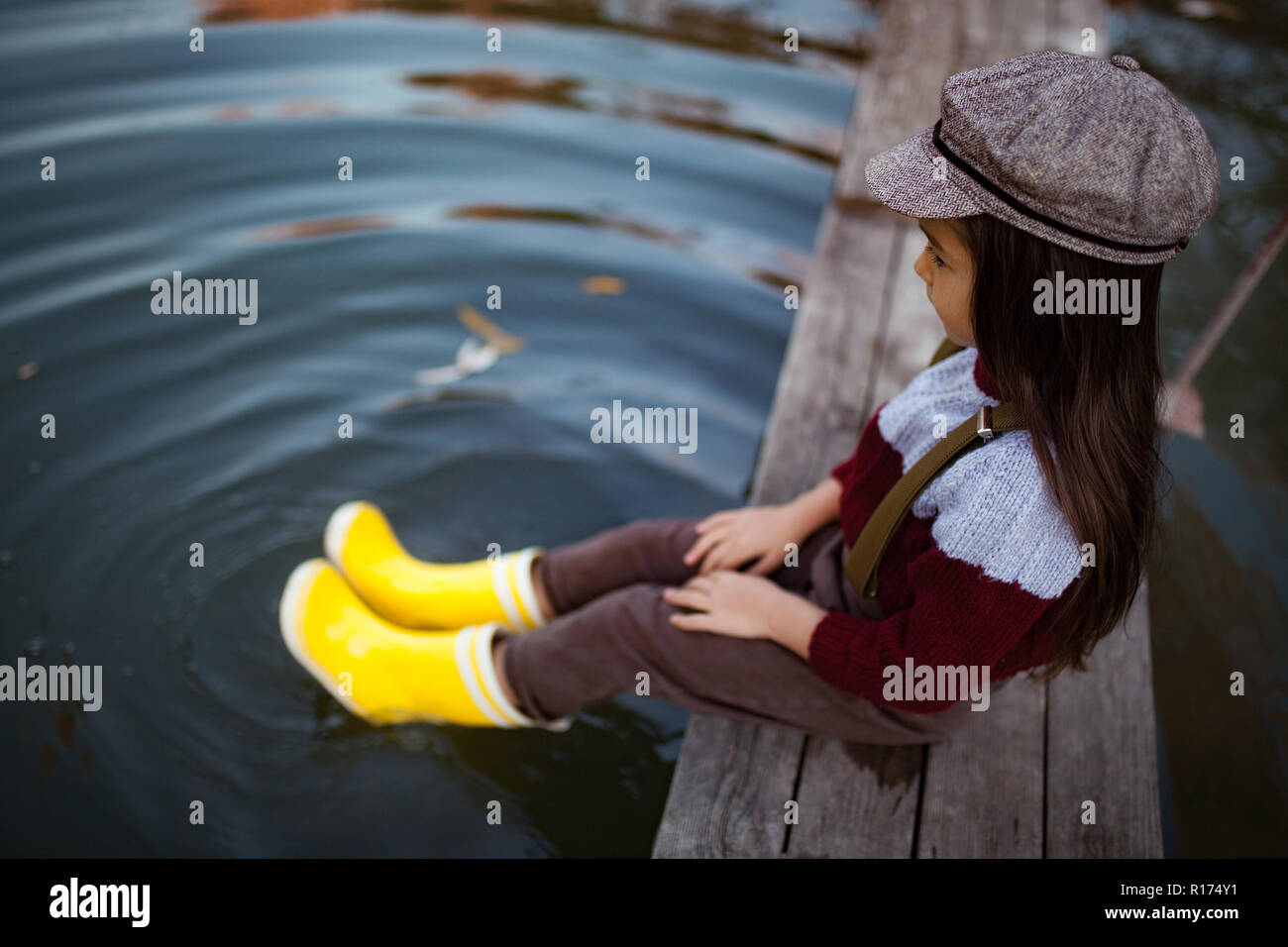 Bambino ragazza in giallo stivali di gomma e cappuccio si siede sul ponte di legno e si mette le gambe in acqua sullo sfondo del fiume. Foto Stock