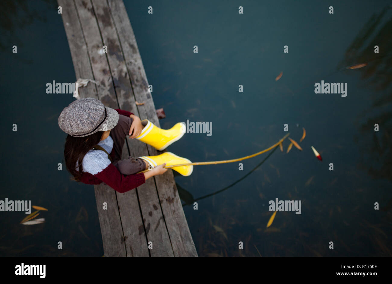 Bambino ragazza in giallo stivali di gomma e cappuccio si siede sul legno ponte di pesca e le catture di pesce con self-made canna da pesca. Vista dall'alto. Foto Stock