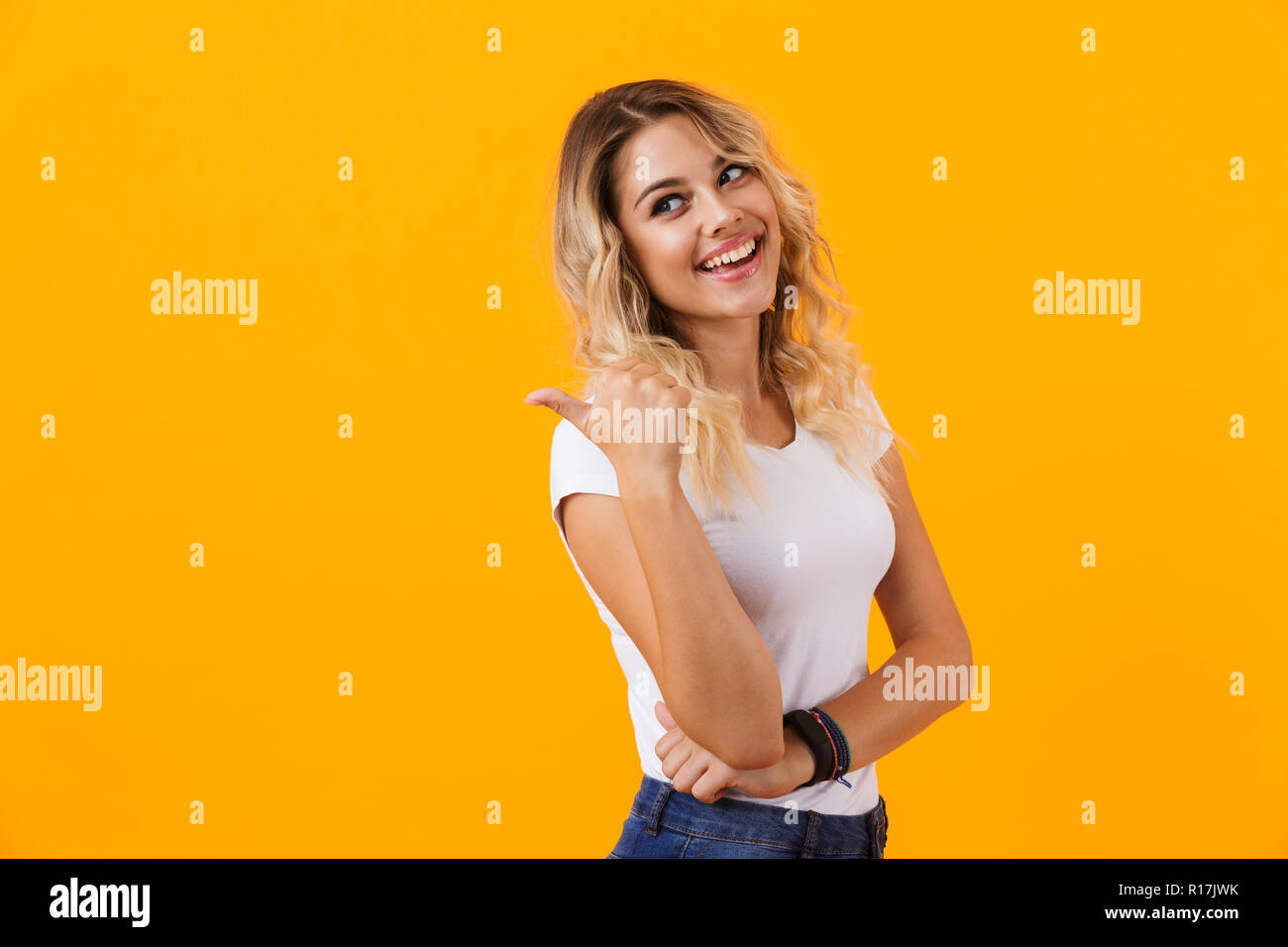 Foto di tipo donna bionda in basic abbigliamento sorridente e puntare il dito a copyspace isolato su sfondo giallo Foto Stock