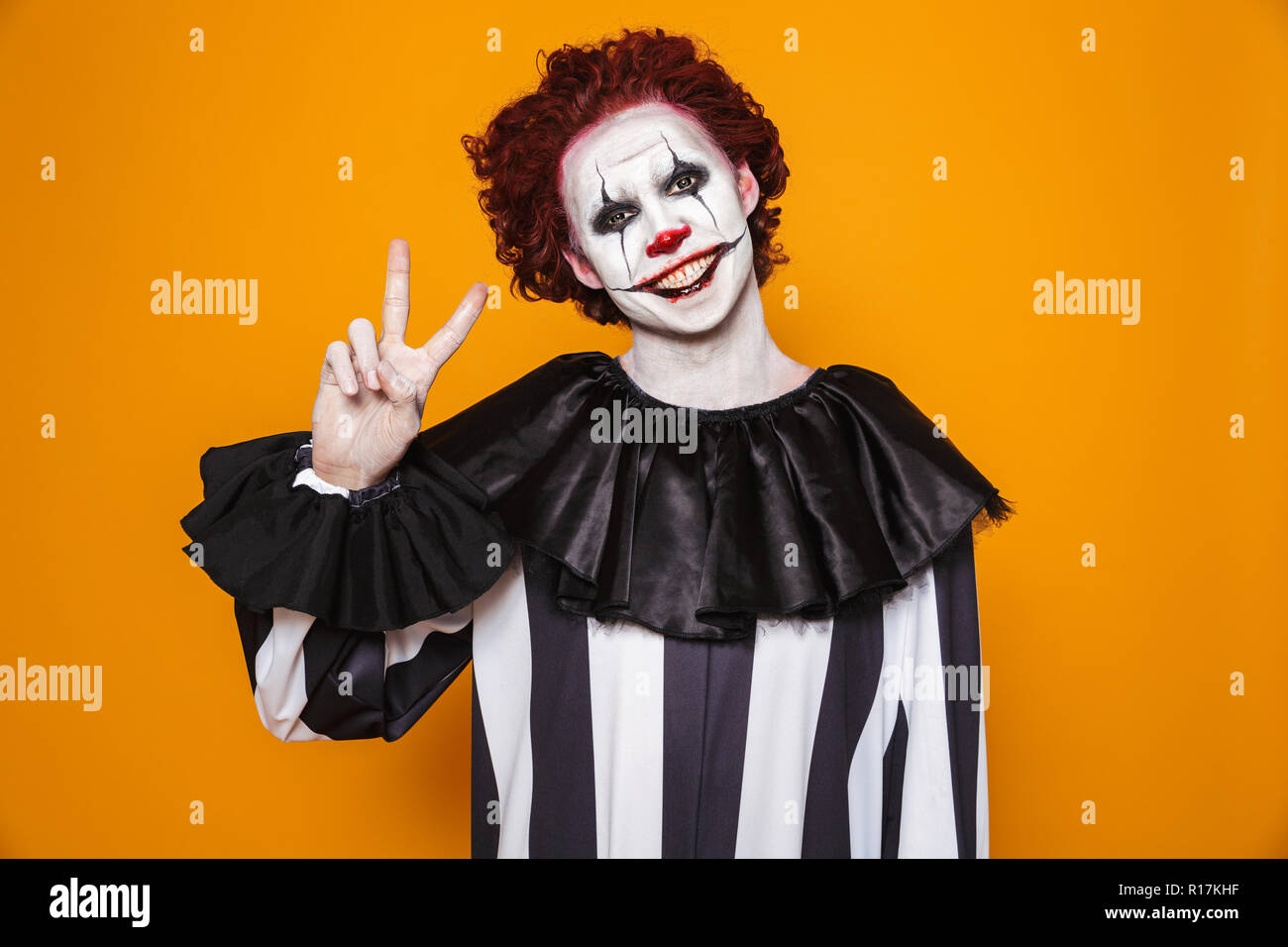 Scary clown uomo 20s indossando il costume nero e Halloween trucco guardando la telecamera isolate su sfondo giallo Foto Stock