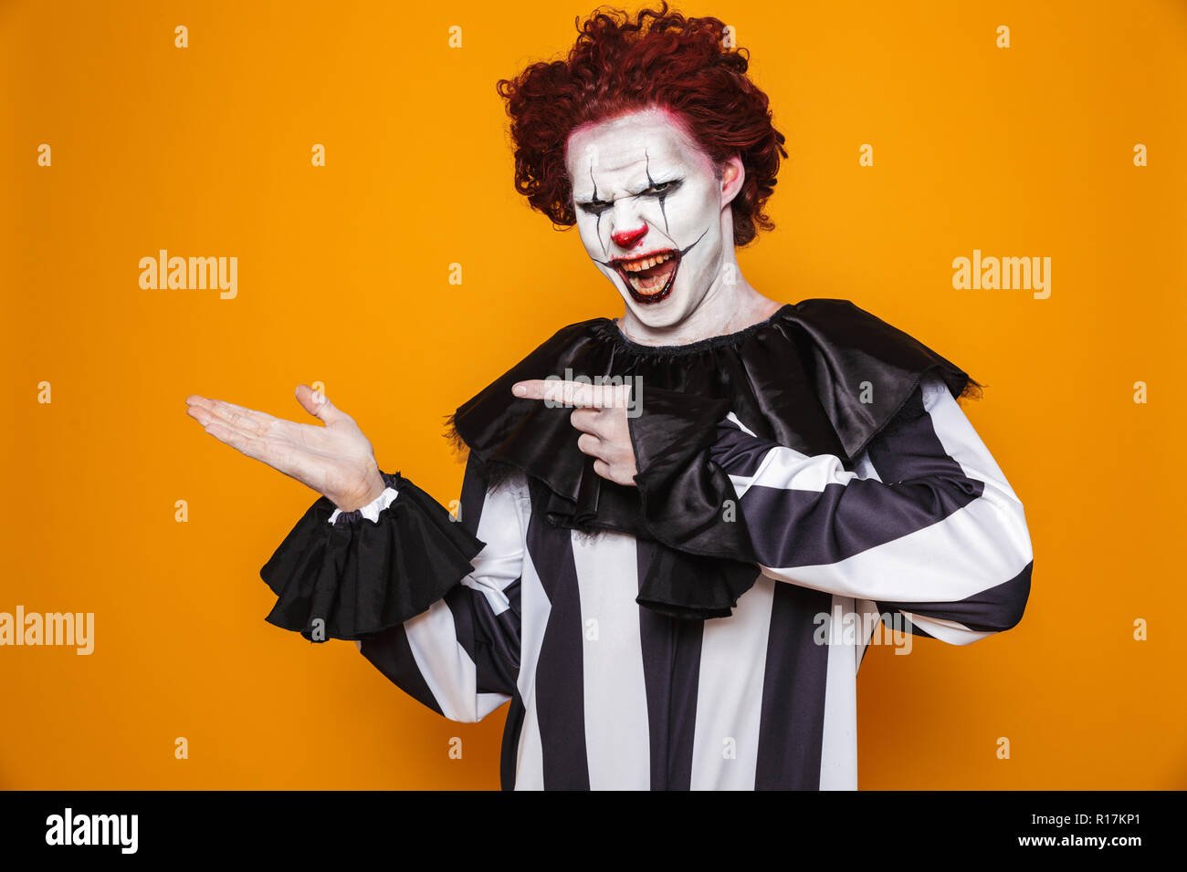 Brutto clown uomo 20s indossando il costume nero e Halloween trucco azienda copyspace presso il palm isolate su sfondo giallo Foto Stock