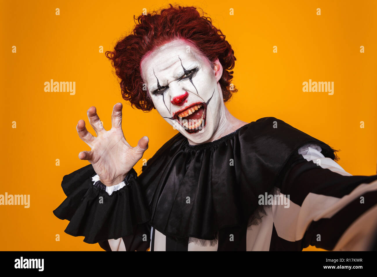 Scioccante clown uomo 20s indossando il costume nero e Halloween trucco smorfie in telecamera isolate su sfondo giallo Foto Stock