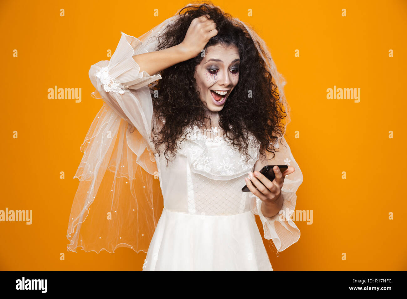 Sorpreso felice sposa zombie in abito da sposa utilizza lo smartphone e sorridente isolate su Orange Foto Stock