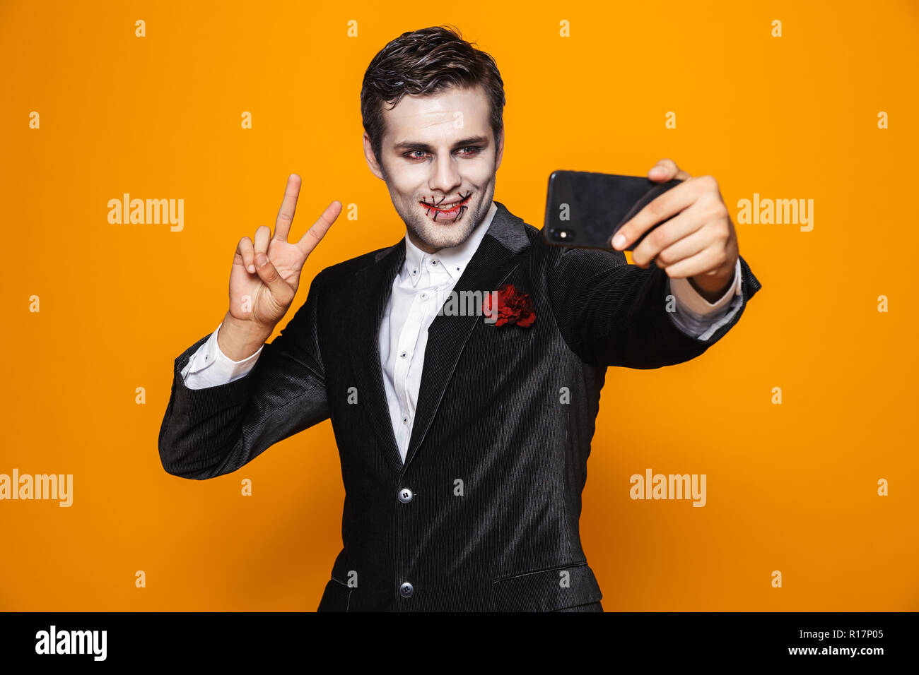 Sorridente uomo bello zombie con halloween make-up che mostra il gesto di pace e rendere selfie sullo smartphone isolate su Orange Foto Stock