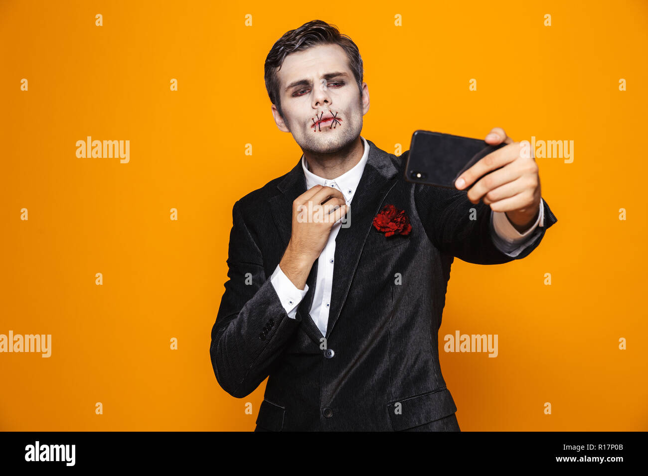 Bel uomo zombie con halloween make-up e in tuta rendendo selfie sullo smartphone isolate su Orange Foto Stock