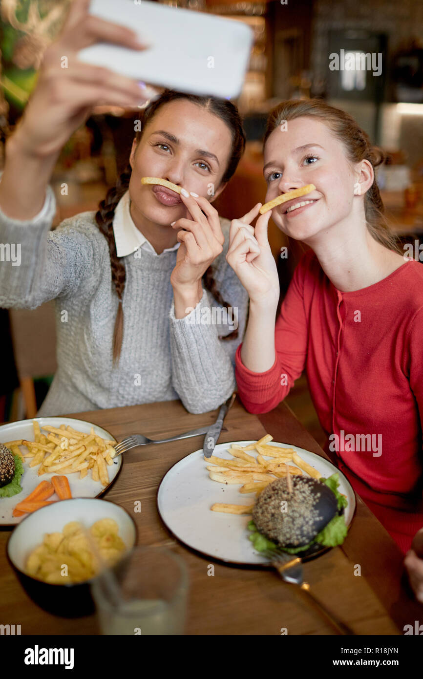 Due ragazze divertente rendendo selfie con patatine fritte tra il naso e le labbra mentre di trascorrere del tempo in un fast food cafe Foto Stock