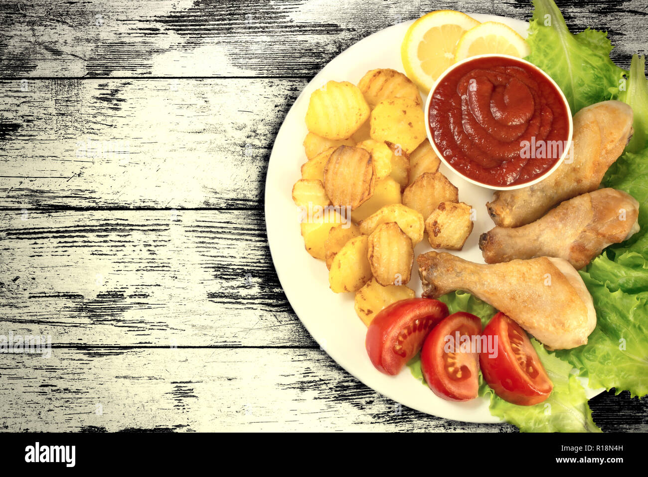 Cosce di pollo su una piastra bianca con fette di pomodoro e insalata e patatine fritte e ketchup sulla tavola di legno tabella sfondo vintage. Vista dall'alto. Vecchio styl Foto Stock