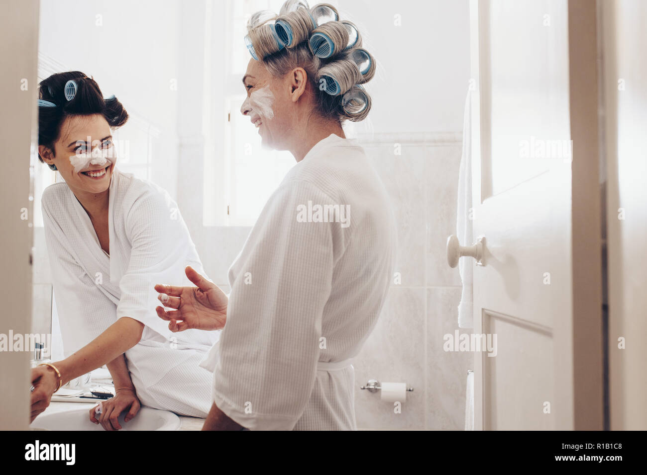 Felice madre e figlia in piedi in bagno accappatoi da indossare con rulli di arricciatura sulla testa. Le donne l'applicazione di crema per il viso in piedi in bagno a h Foto Stock