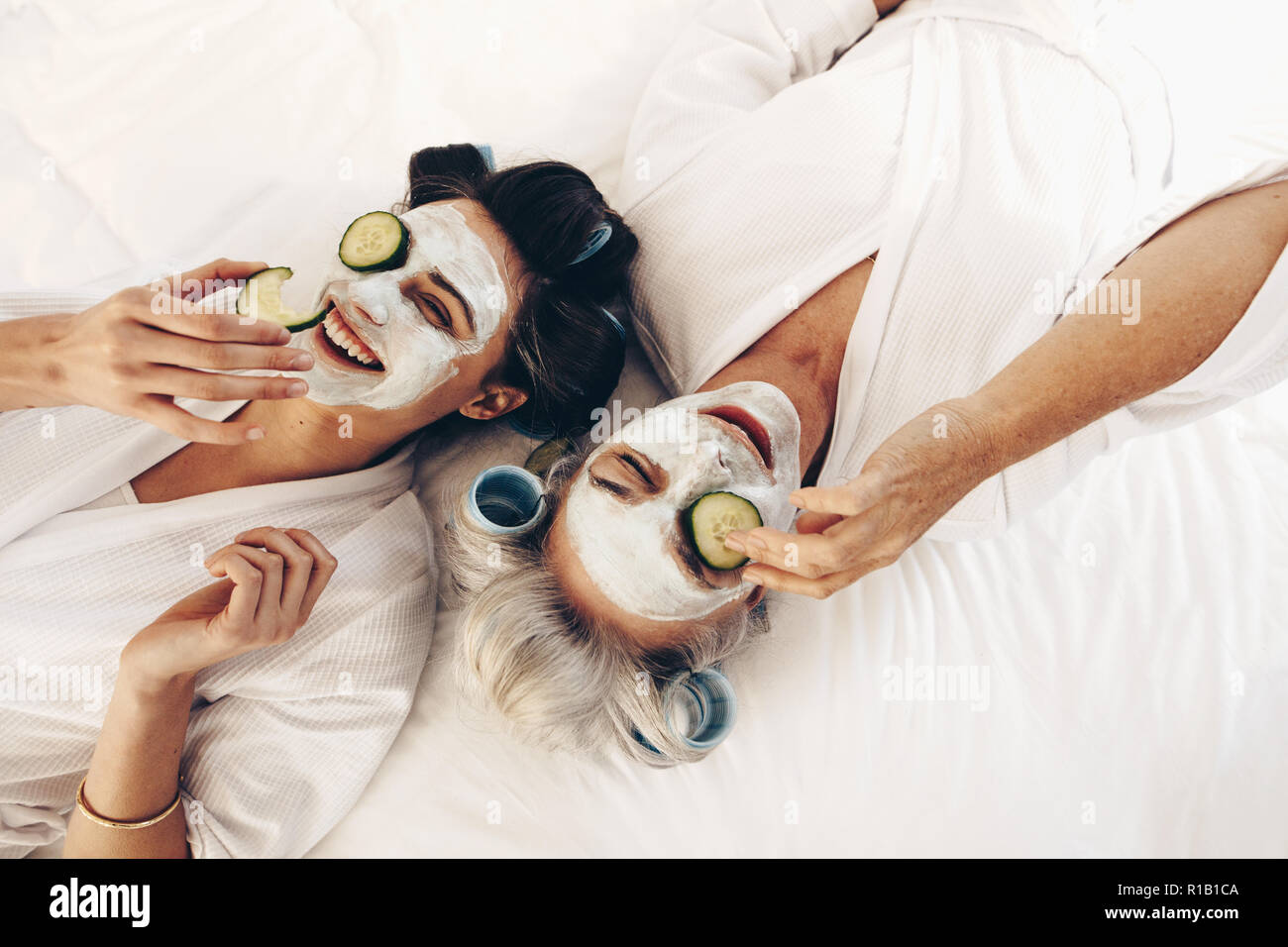 Due donne in accappatoi giacente sul letto indossando bellezza viso pack con fette di cetriolo. Madre e figlia rilassante sul letto indossando face pack e rullo di cu Foto Stock