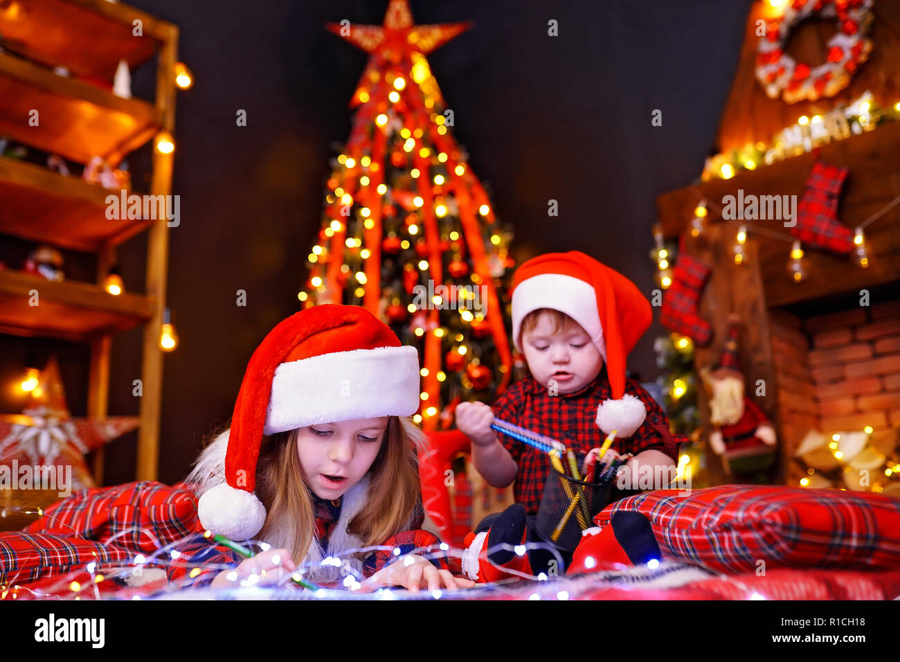 Affascinante ragazza in Santa hat sdraiato sul pavimento e scrive la lettera, disegna con la matita e il suo piccolo fratello che interferisce con la sua, gioca con matite in camera calda con ghirlande, luci Foto Stock