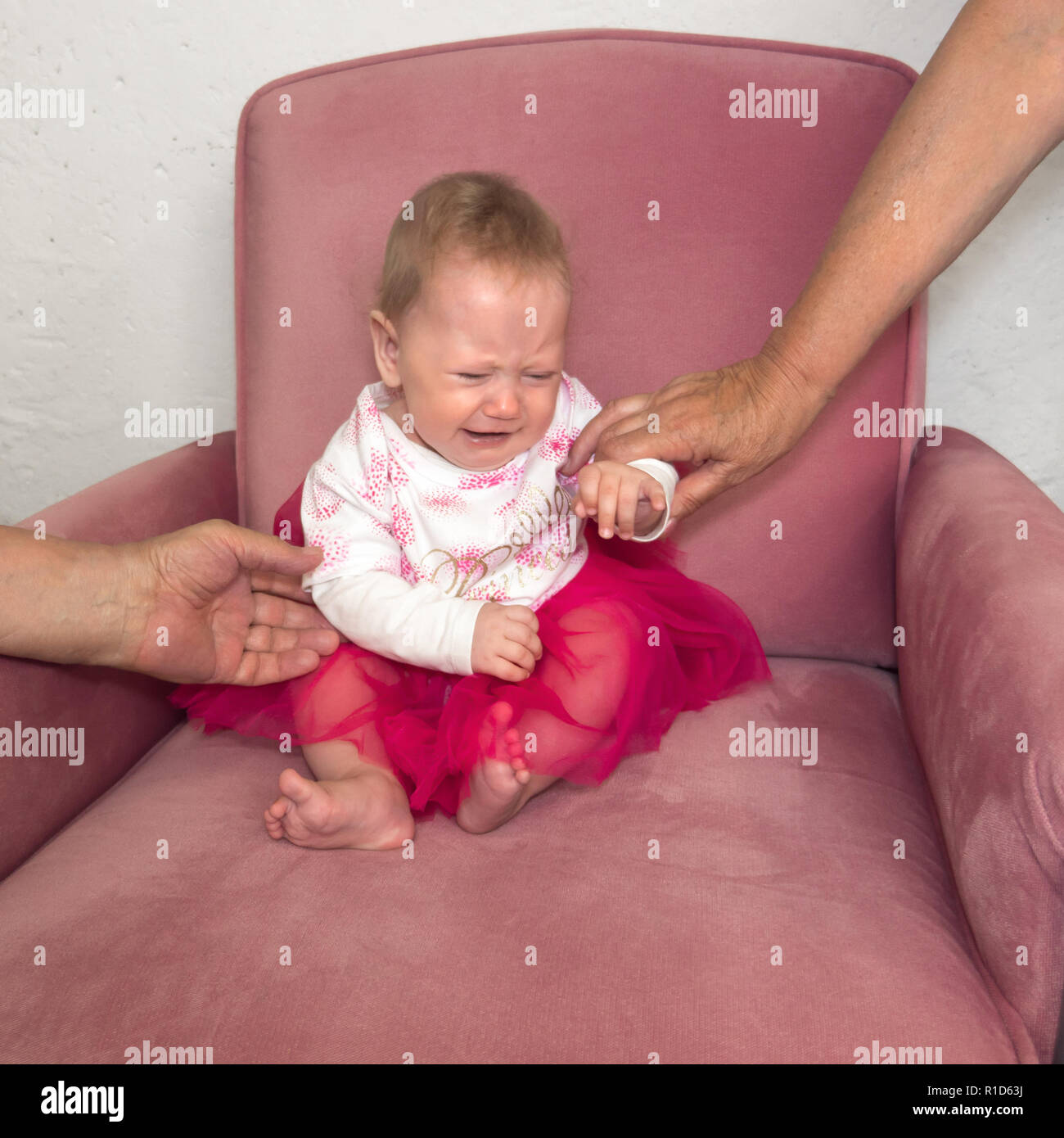 Gridando bambino seduto in poltrona. Le mani di nonna holding bambino seduto in poltrona. Bambino negativo emozione. L'espressione del viso. Ritratto di vestito ragazza seduta sulla sedia. Foto Stock