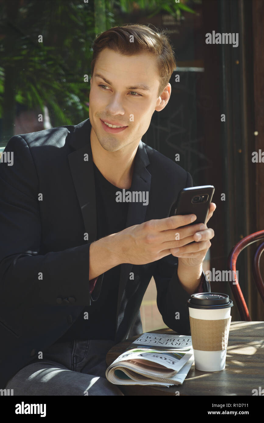 Un giovane imprenditore in possesso di un cellulare,seduta outdoor presso una caffetteria, guardando lontano e sorridente. Foto Stock