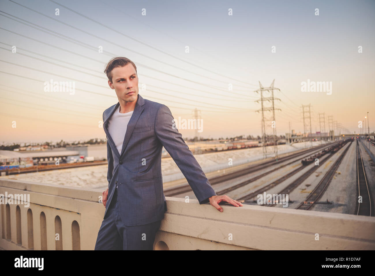 Un bel maschio modello vestito con un abito grigio in piedi da un ponte a Los Angeles al tramonto con uno splendido skyline. Egli guarda lontano e sembra ambizioso. Foto Stock