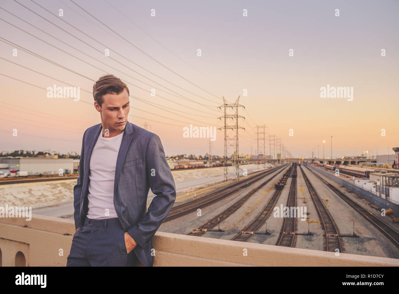 Un bel maschio modello vestito con un abito grigio in piedi da un ponte a Los Angeles al tramonto con uno splendido skyline. Egli guarda giù nel profondo del pensiero. Foto Stock