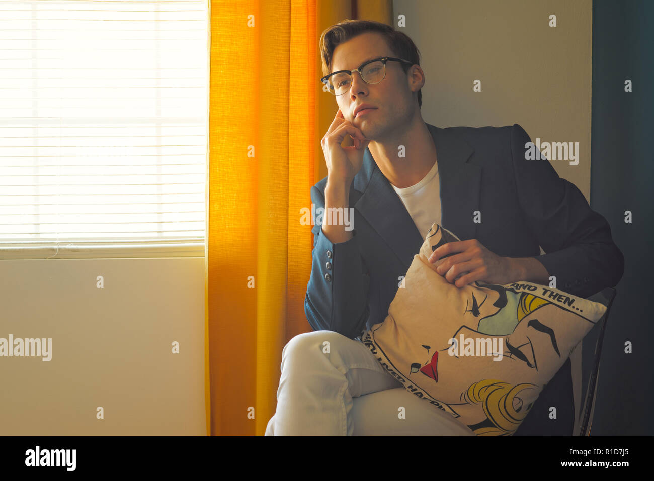 Un giovane uomo, maschio modello in tuta blu e bianco pantaloni,indossa un paio di occhiali vintage, mano al mento tenendo un cuscino. Egli guarda lontano, nel profondo del pensiero. Foto Stock