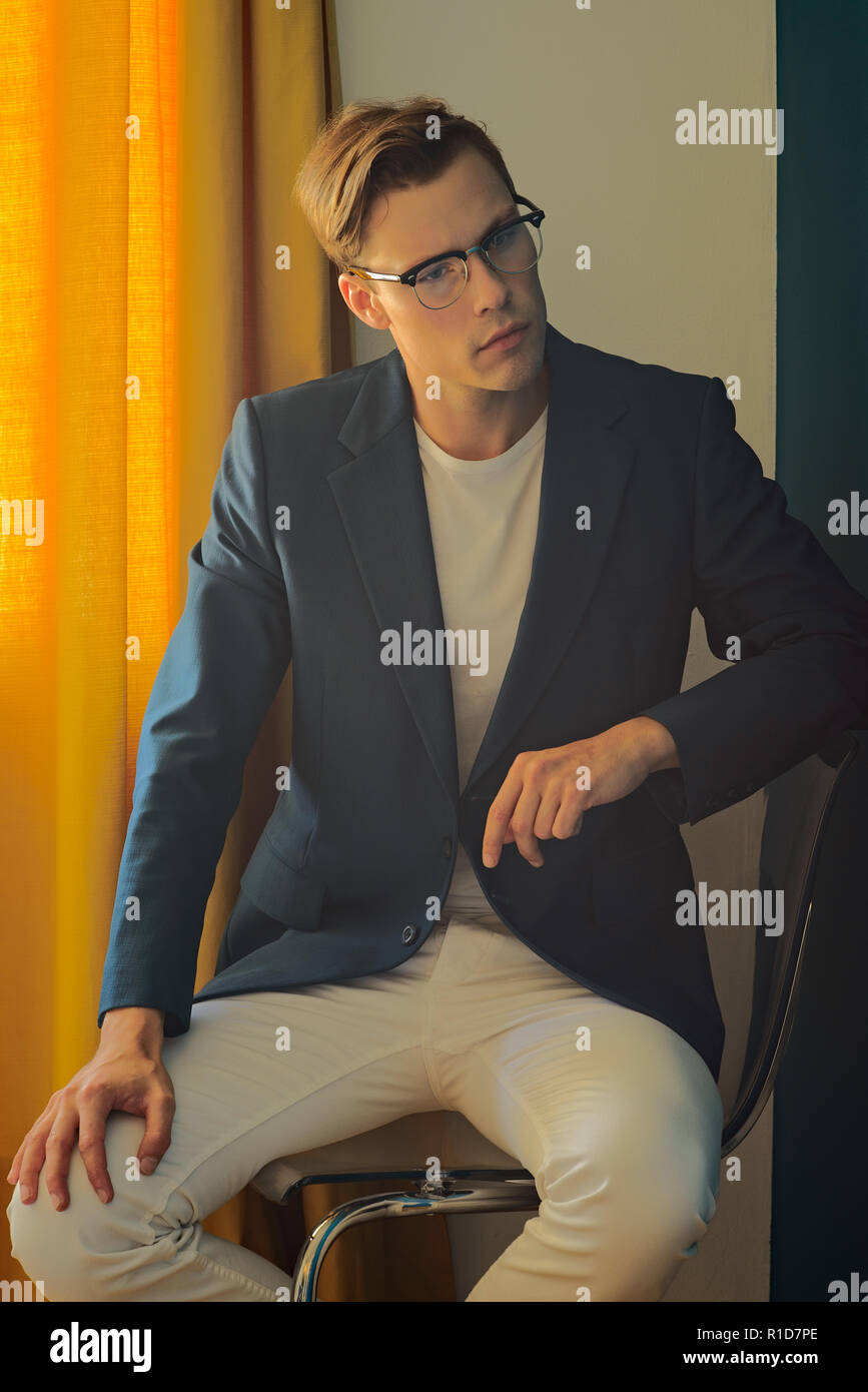 Un giovane uomo, maschio modello in tuta blu e bianco pantaloni,indossa un paio di occhiali vintage, seduto e guardando lontano, nel profondo del pensiero. Foto Stock