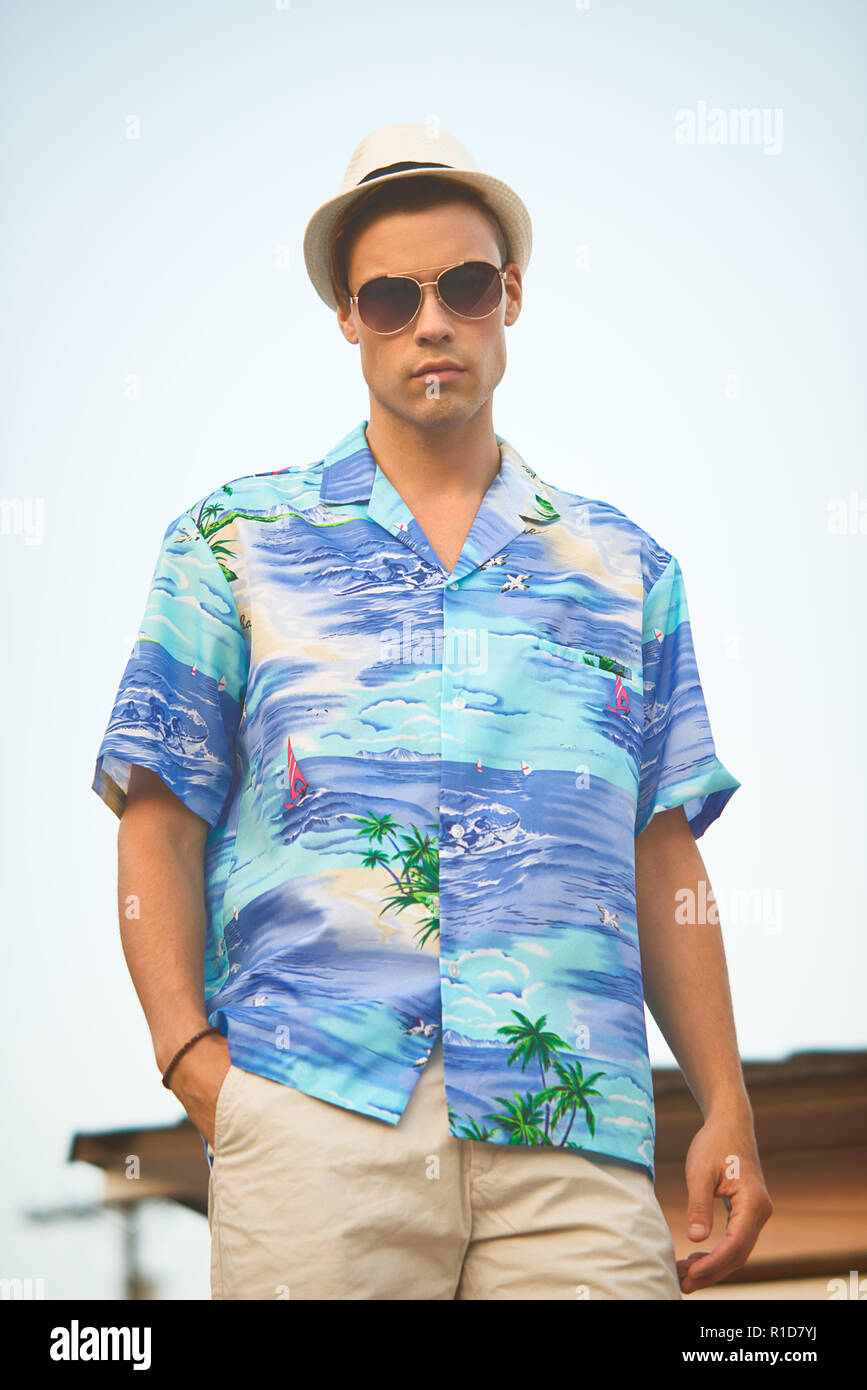 Un bel modello maschile che pongono in estate shirt outdoor contro un cielo blu. Egli indossa una veste bianca cappello Fedora e aviator occhiali da sole. Foto Stock