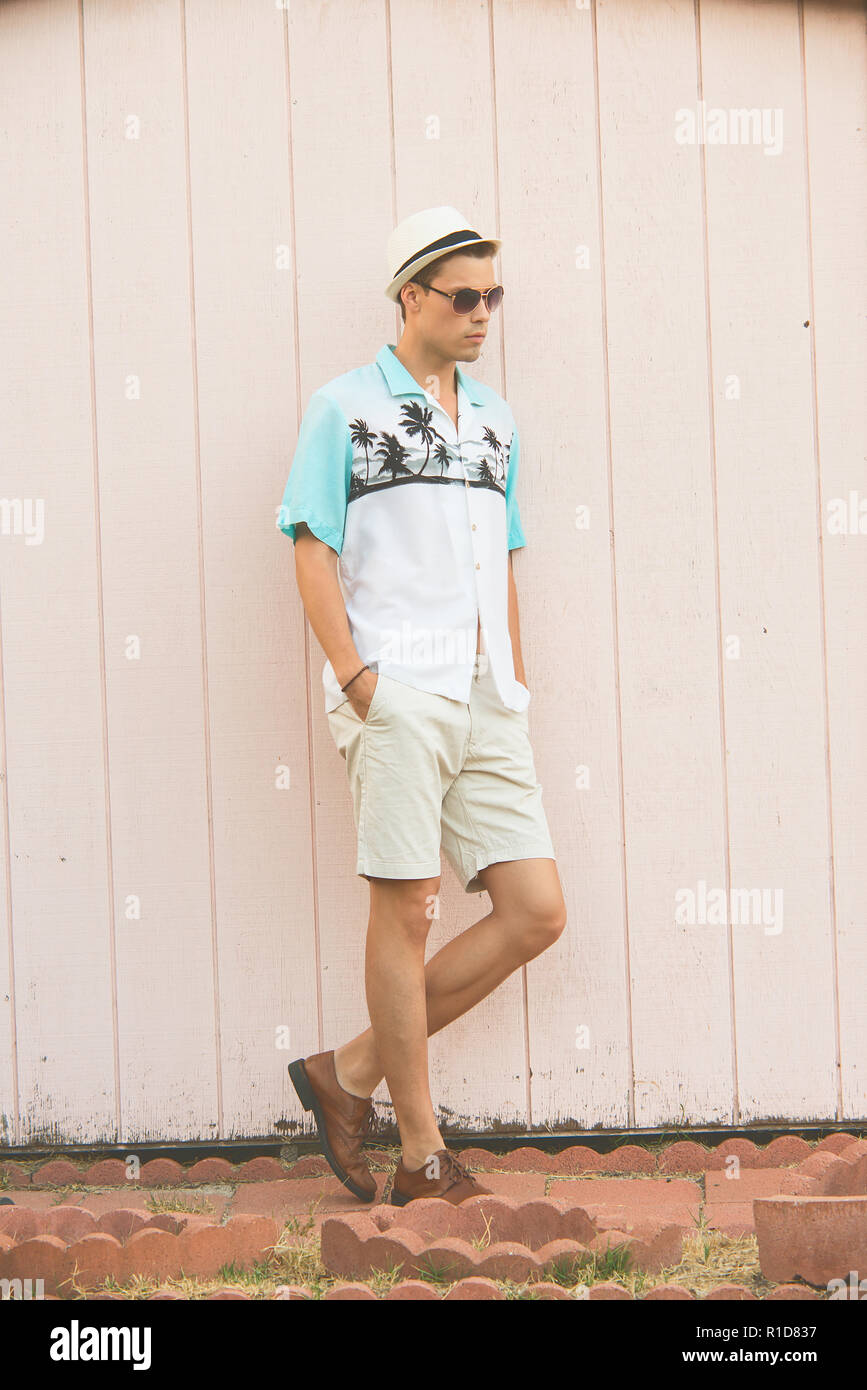 Un bel modello maschile in posa di vestiti estivi all'aperto in piedi contro una normale parete in legno. Foto Stock
