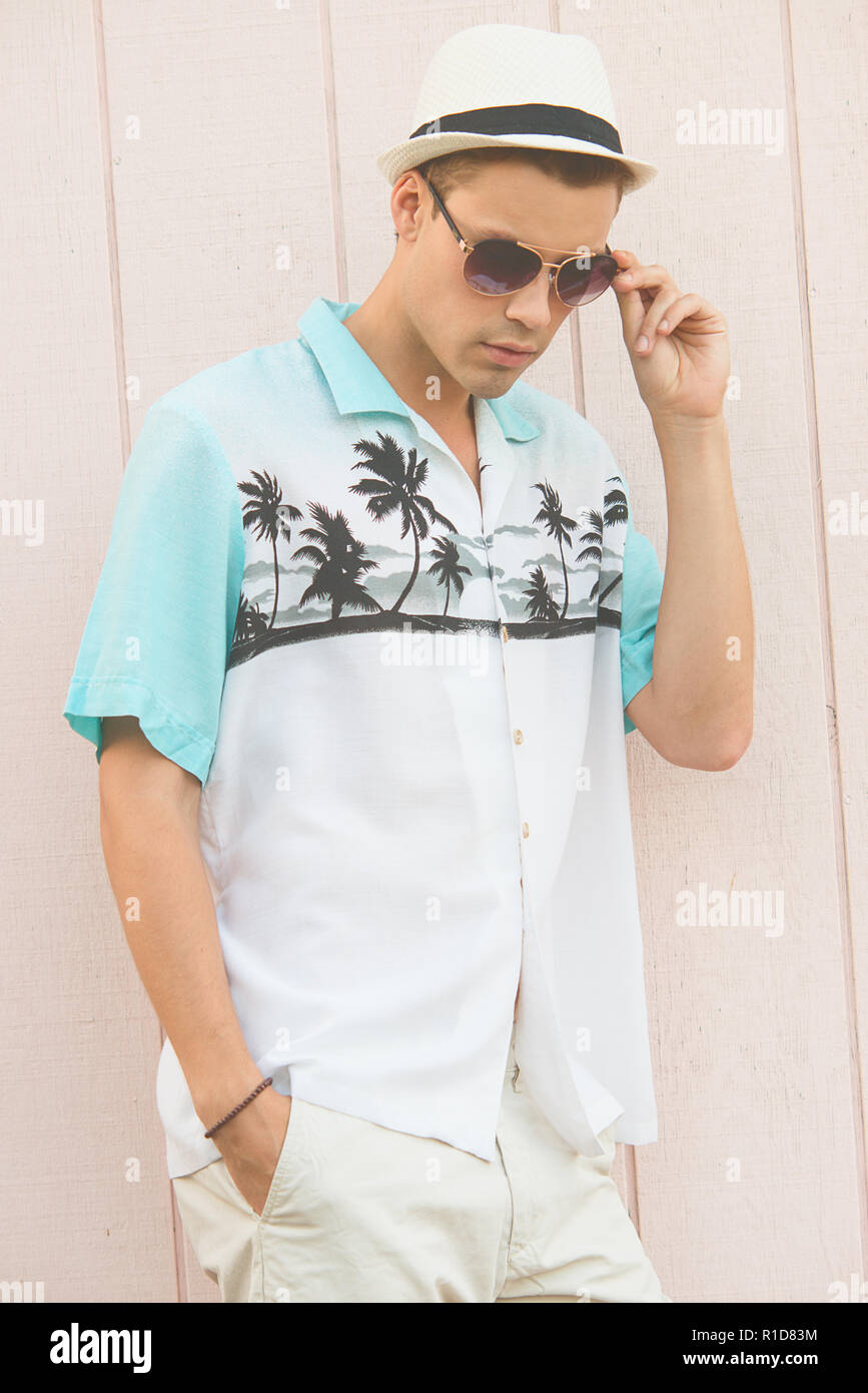 Un bel modello maschile in posa di vestiti estivi all'aperto contro una normale parete in legno. Egli indossa una veste bianca cappello Fedora e aviator occhiali da sole. Foto Stock