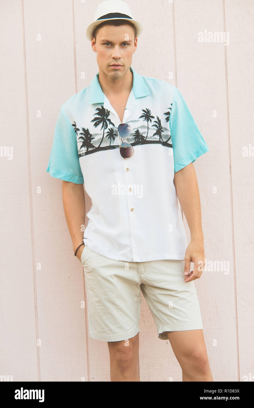 Un bel modello maschile in posa di vestiti estivi all'aperto contro una normale parete in legno. Egli indossa una veste bianca cappello Fedora, guardando fiduciosi. Foto Stock