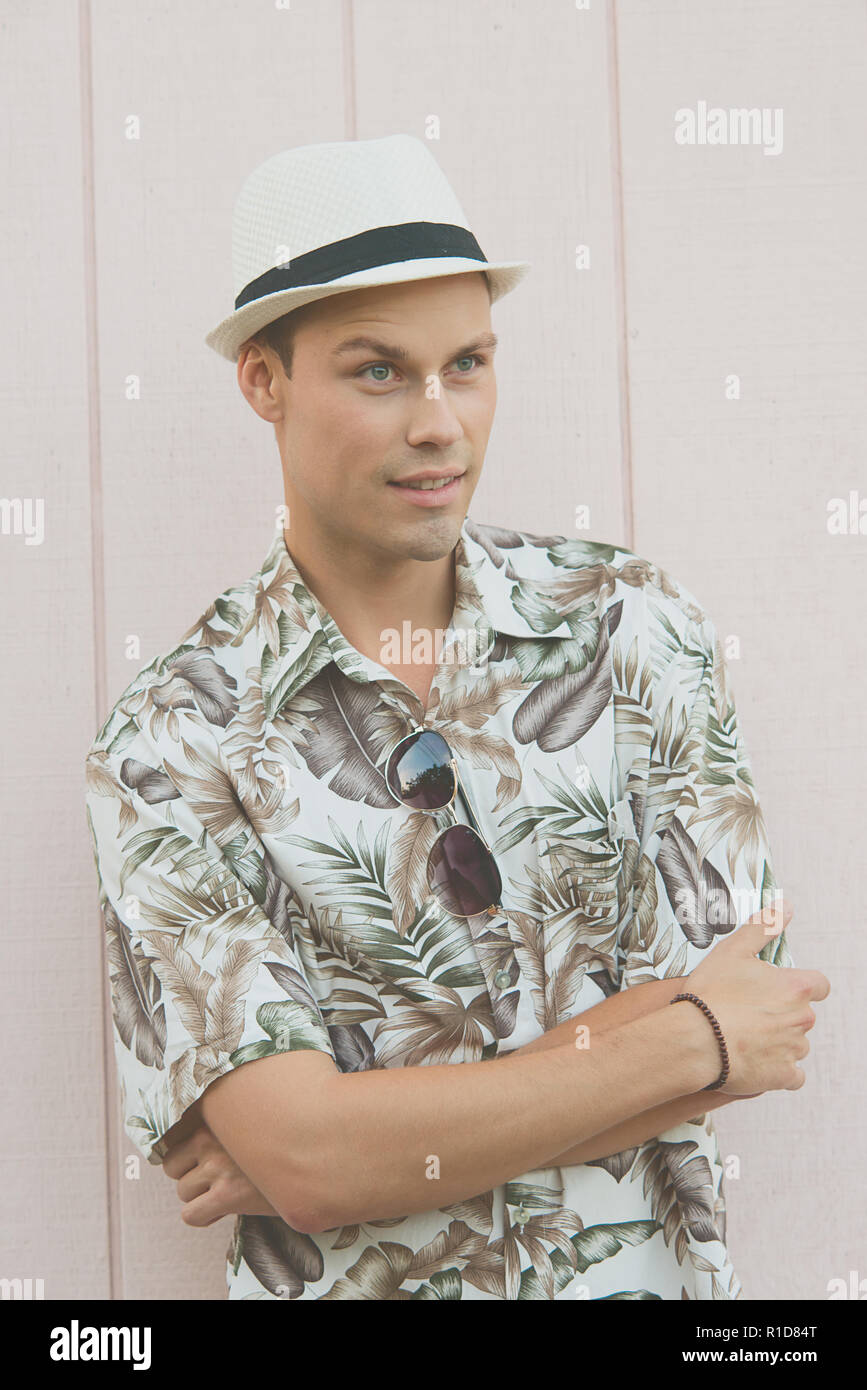 Un bel modello maschile in posa di vestiti estivi all'aperto di fronte ad una normale parete in legno. Indossa un cappello bianco, sembra facile andare e rilassato. Foto Stock