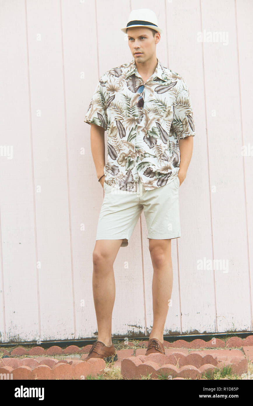 Un uomo bello, modello maschile in posa di vestiti estivi all'aperto in piedi contro una normale parete in legno. Indossa un cappello bianco. Foto Stock