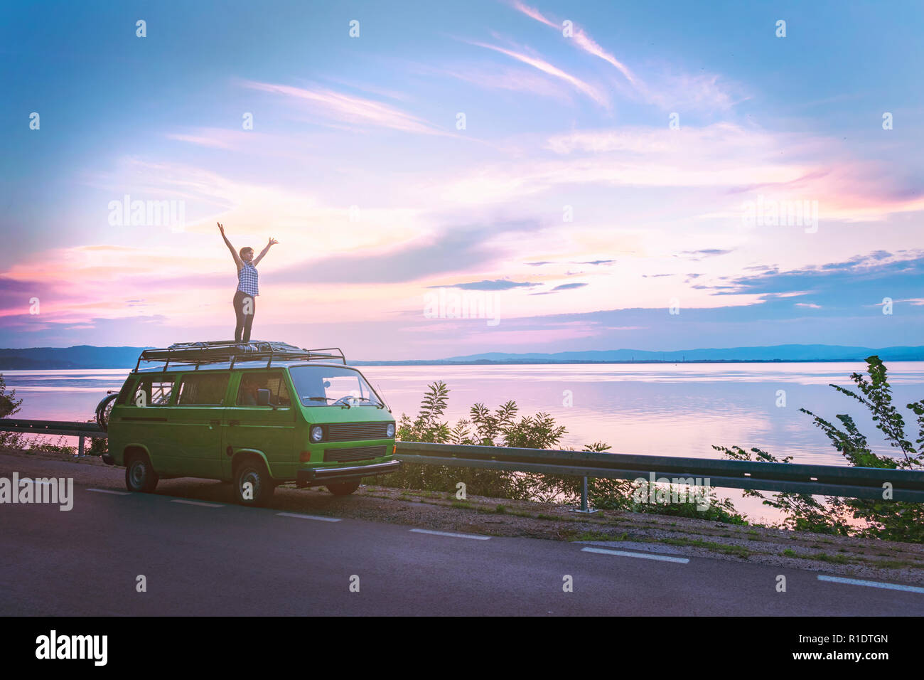 Giovane bella ragazza in piedi sul tetto del vecchio classico timer camper parcheggiato sul mare con incredibilmente colorato tramonto Cielo, mani spalancate libera Foto Stock