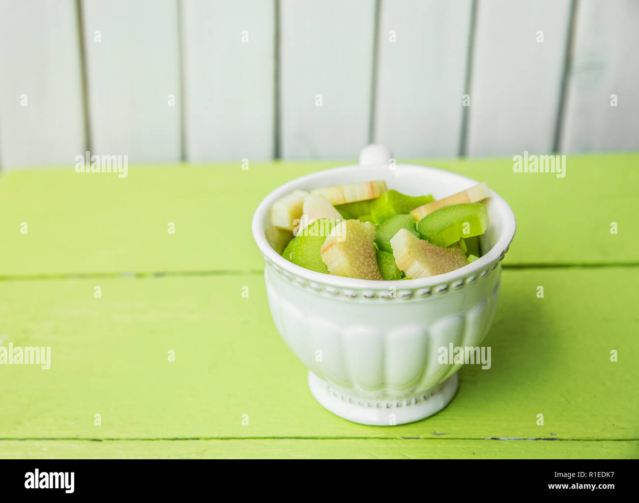 Fresche e mature di rabarbaro (Rheum rhabarbarum) sbucciato e tagliato a pezzi in un bianco vintage cup. Preparati per la cottura. Verde e bianco sullo sfondo di legno. Foto Stock