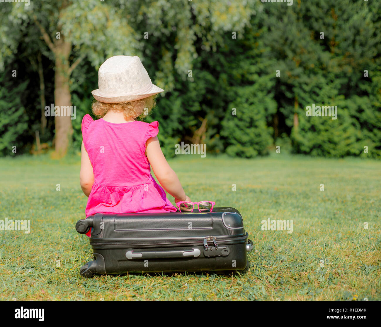 Vista dal retro, anonimi ragazza giovane bambino in abito rosa si siede su un nero per i bagagli a mano e attende, parco all'aperto in estate. Foto Stock