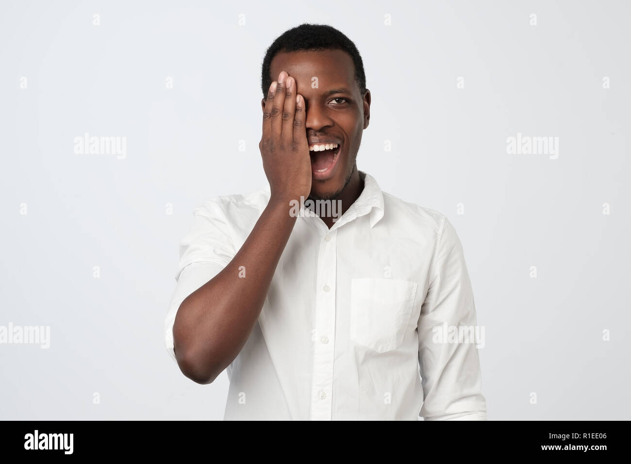 I giovani africani Uomo in camicia bianca che copre metà faccia con la mano mentre in piedi contro uno sfondo grigio. Positivo emozione umana Foto Stock