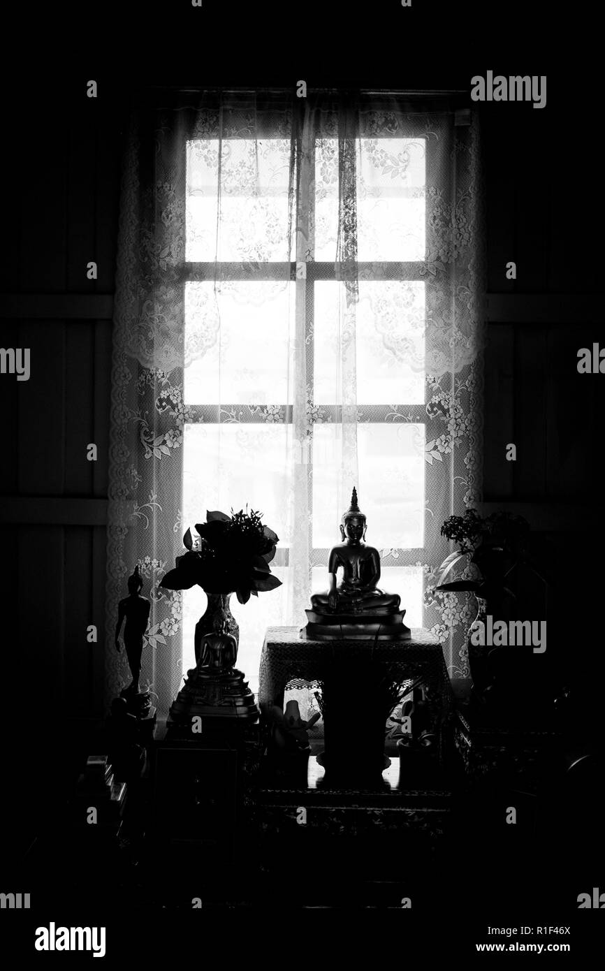 Quasi silhouette oro statue di Buddha seduto presso la finestra con tenda bianca. Thailandia santuario domestico. in bianco e nero il filtro. Foto Stock