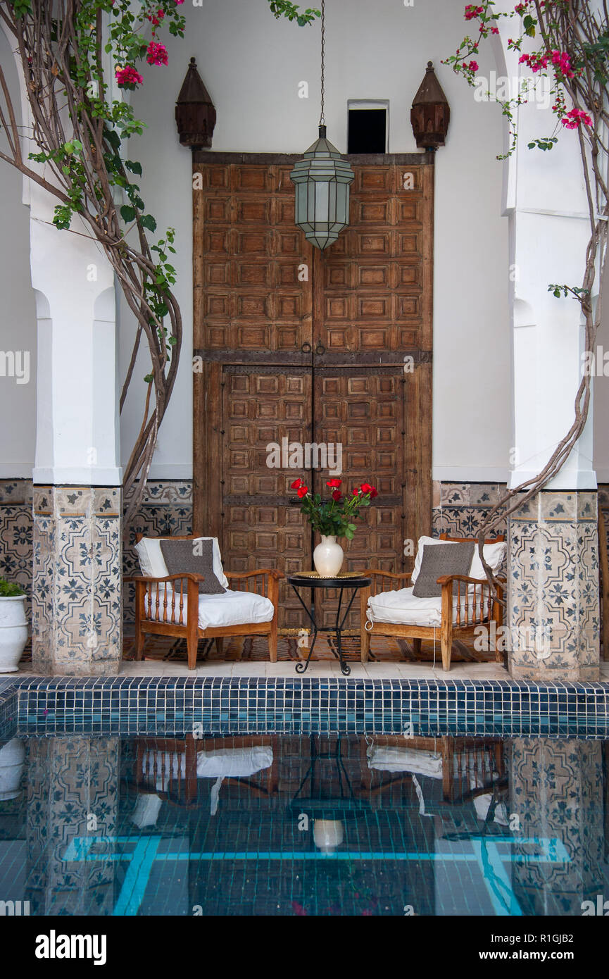 18-04-11. Marrakech, Marocco. Tipico riad marocchino courtyars con alte porte scolpite, piscina di raffreddamento, piscina, sedie e rose in un vaso. Foto Foto Stock