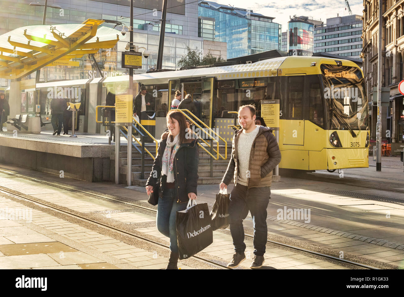 2 Novembre 2018: Manchester, Regno Unito - coppia giovane e sorridente con Debenhams borse per lo shopping in Exchange Square, con un tram Metrolink dietro. Un sacco di flare. Foto Stock