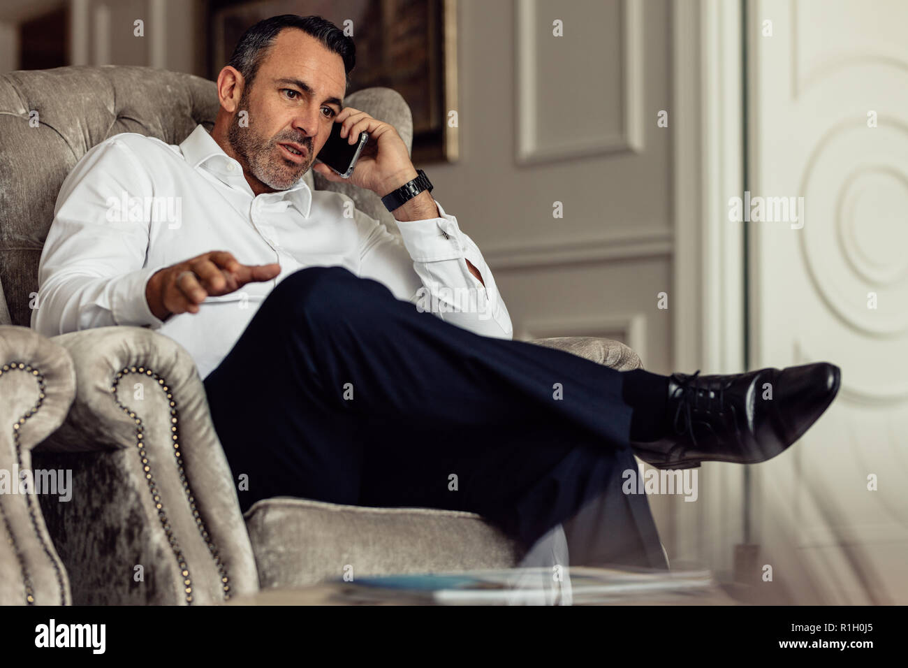 Viaggiatore maturo uomo d'affari che indossa una camicia bianca che effettua una chiamata dopo l'arrivo in camera. viaggiatore seduto sul divano e a parlare sullo smartphone. Foto Stock