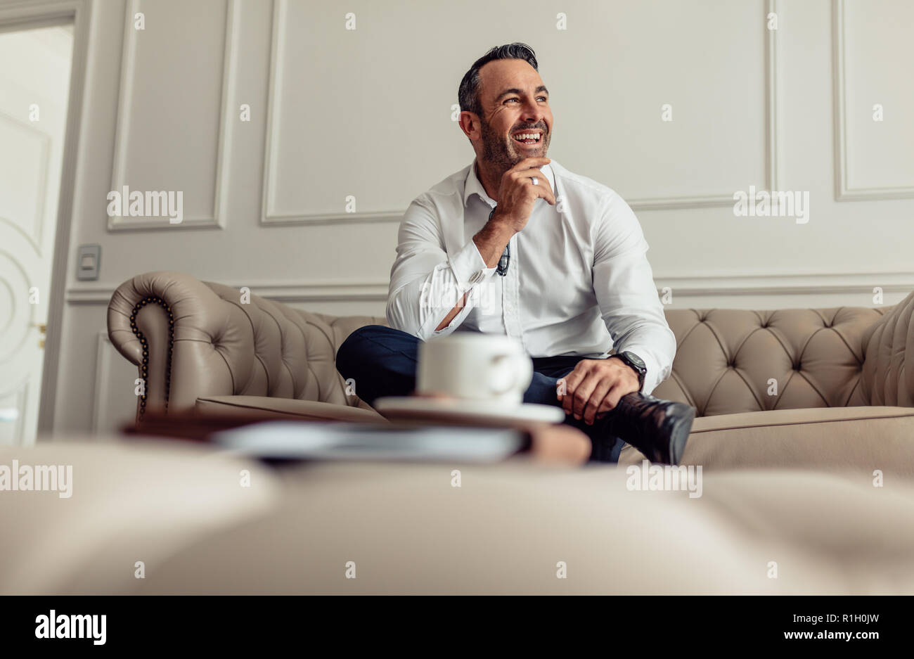 Ritratto di imprenditore sorridente seduto sul divano nella camera di albergo e guardando lontano. Imprenditore maturo alloggiare in un lussuoso hotel room in viaggio d'affari. Foto Stock