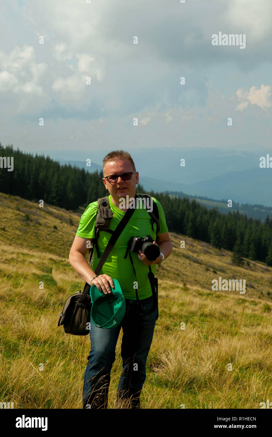 Описаphotographer in un verde T-shirt e jeans blu su una luminosa giornata soleggiata con una telecamera e un cappuccio verde nelle sue mani nelle montagne di fronte al clo Foto Stock