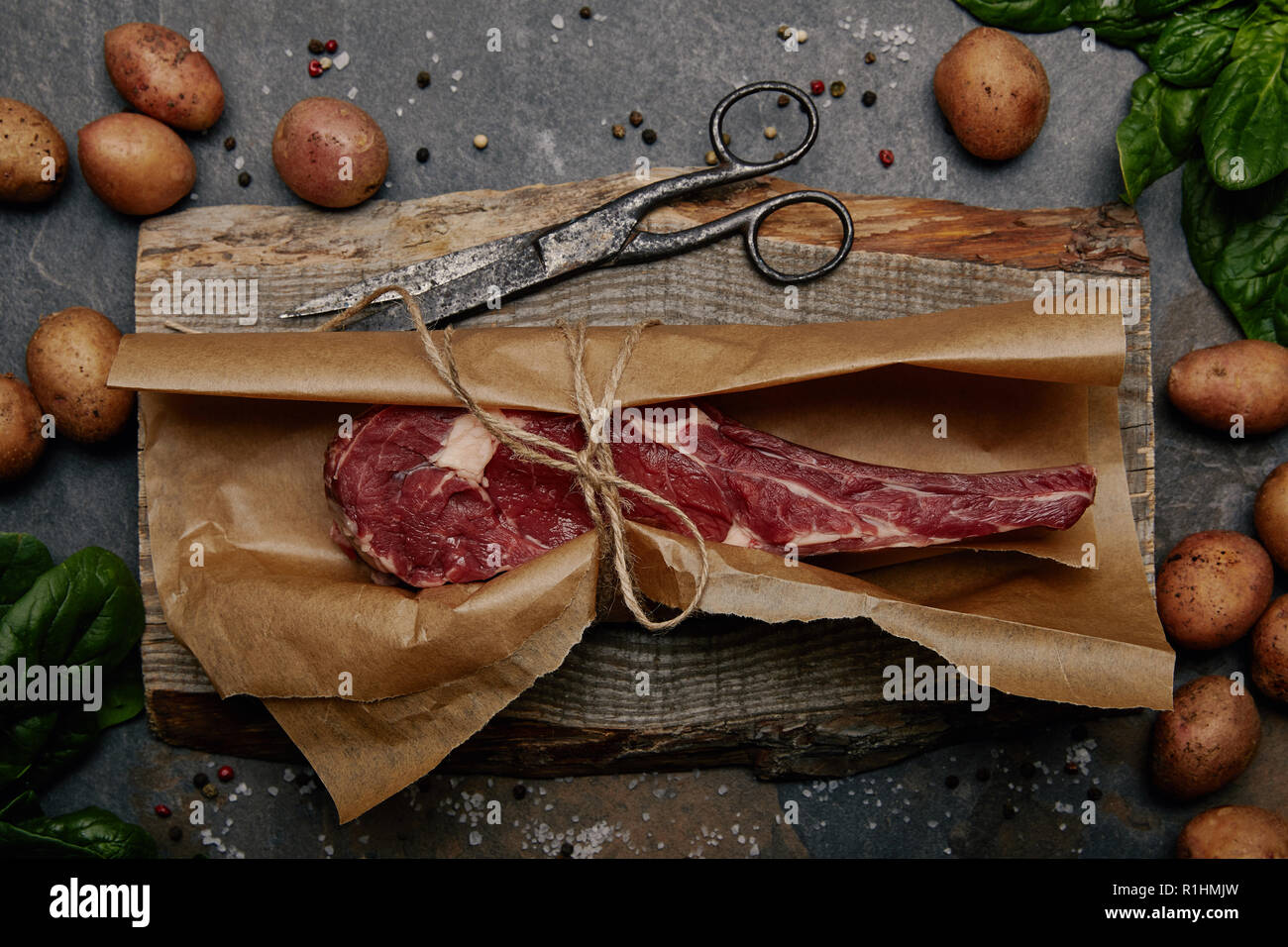 Vista superiore della nervatura di greggio eye steak avvolto in carta da forno su tavola di legno con spezie e patate Foto Stock