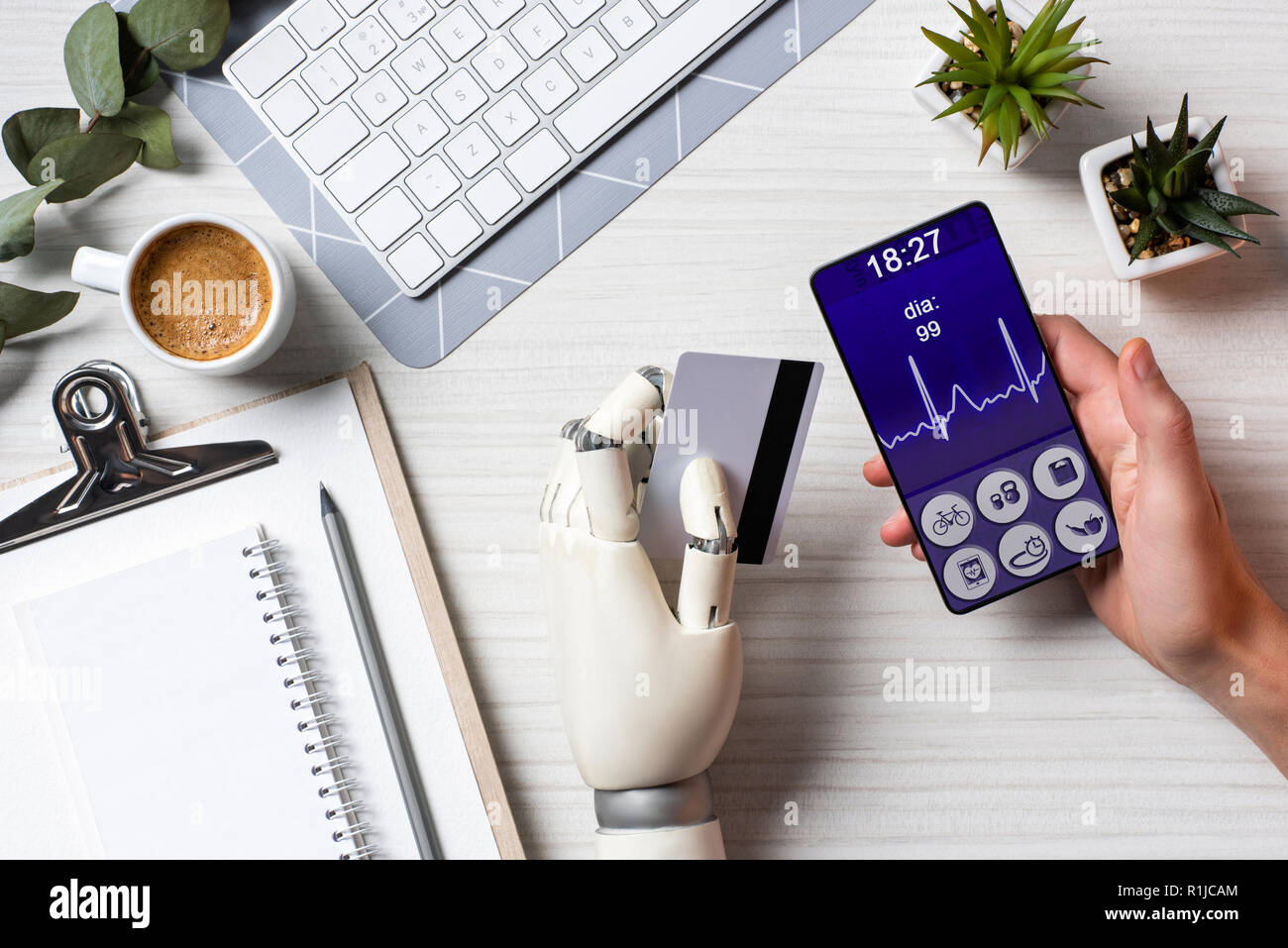 Vista parziale di imprenditore cyborg con la mano che tiene la carta di credito e utilizza lo smartphone con applicazione medica sullo schermo a tavola con la tazza di caffè in o Foto Stock