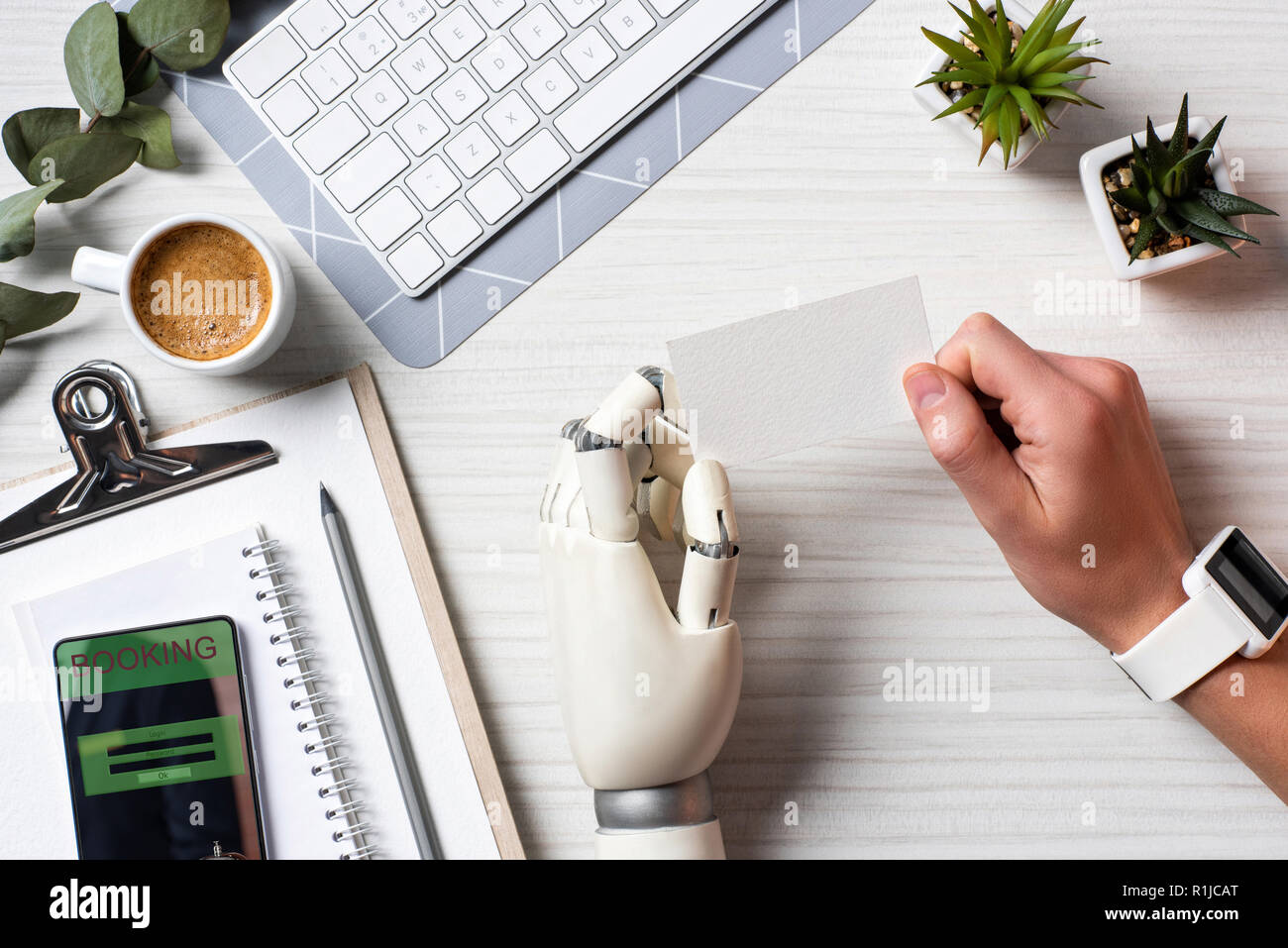 Immagine ritagliata di imprenditore con mano cyborg e smartwatch holding blank carta da visita a tavola con lo smartphone con prenotazione sullo schermo in office Foto Stock