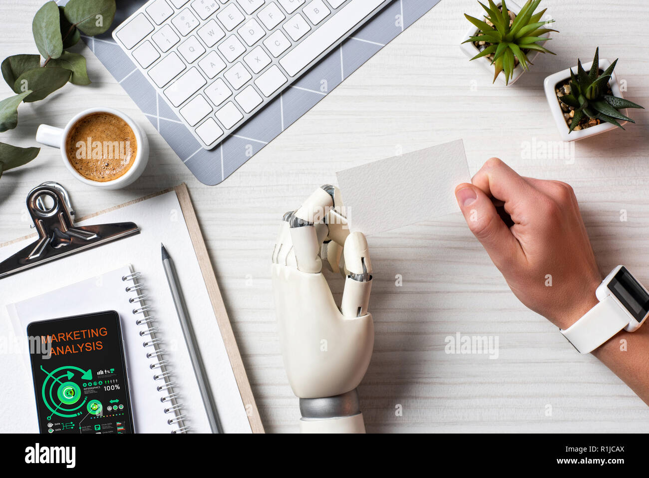 Vista parziale di imprenditore con mano cyborg e smartwatch holding blank carta da visita a tavola con lo smartphone con analisi di marketing sullo schermo in off Foto Stock