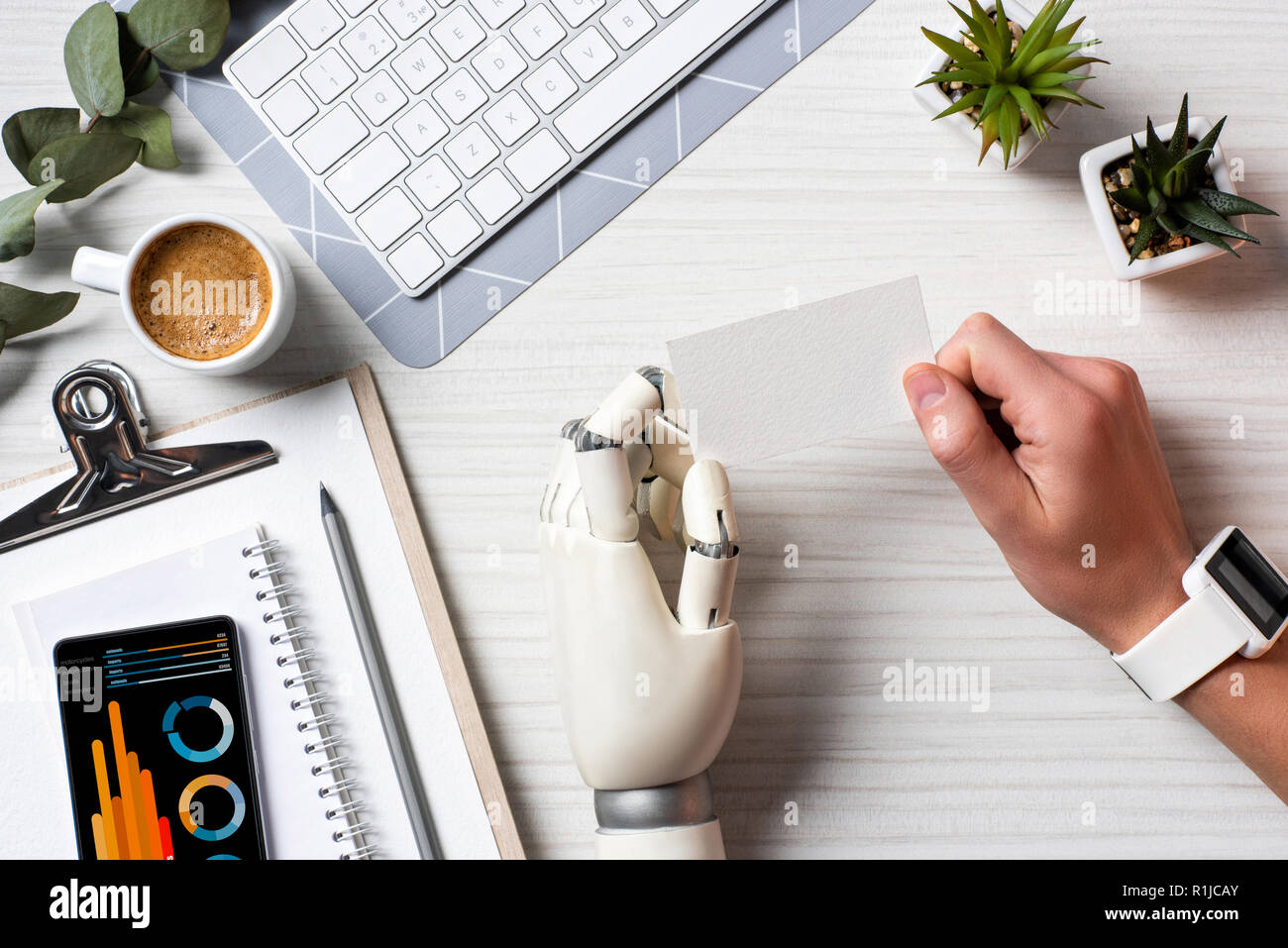 Immagine ritagliata di imprenditore con mano cyborg e smartwatch holding blank carta da visita a tavola con lo smartphone con una infografica sullo schermo in office Foto Stock