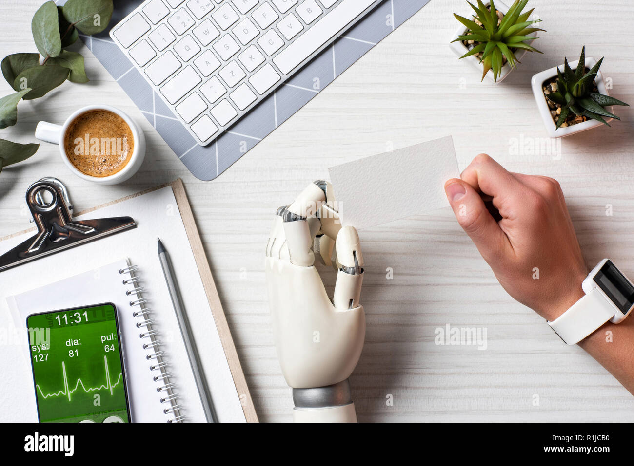Immagine ritagliata di imprenditore con mano cyborg e smartwatch holding blank carta da visita a tavola con lo smartphone con applicazione medica sullo schermo in o Foto Stock