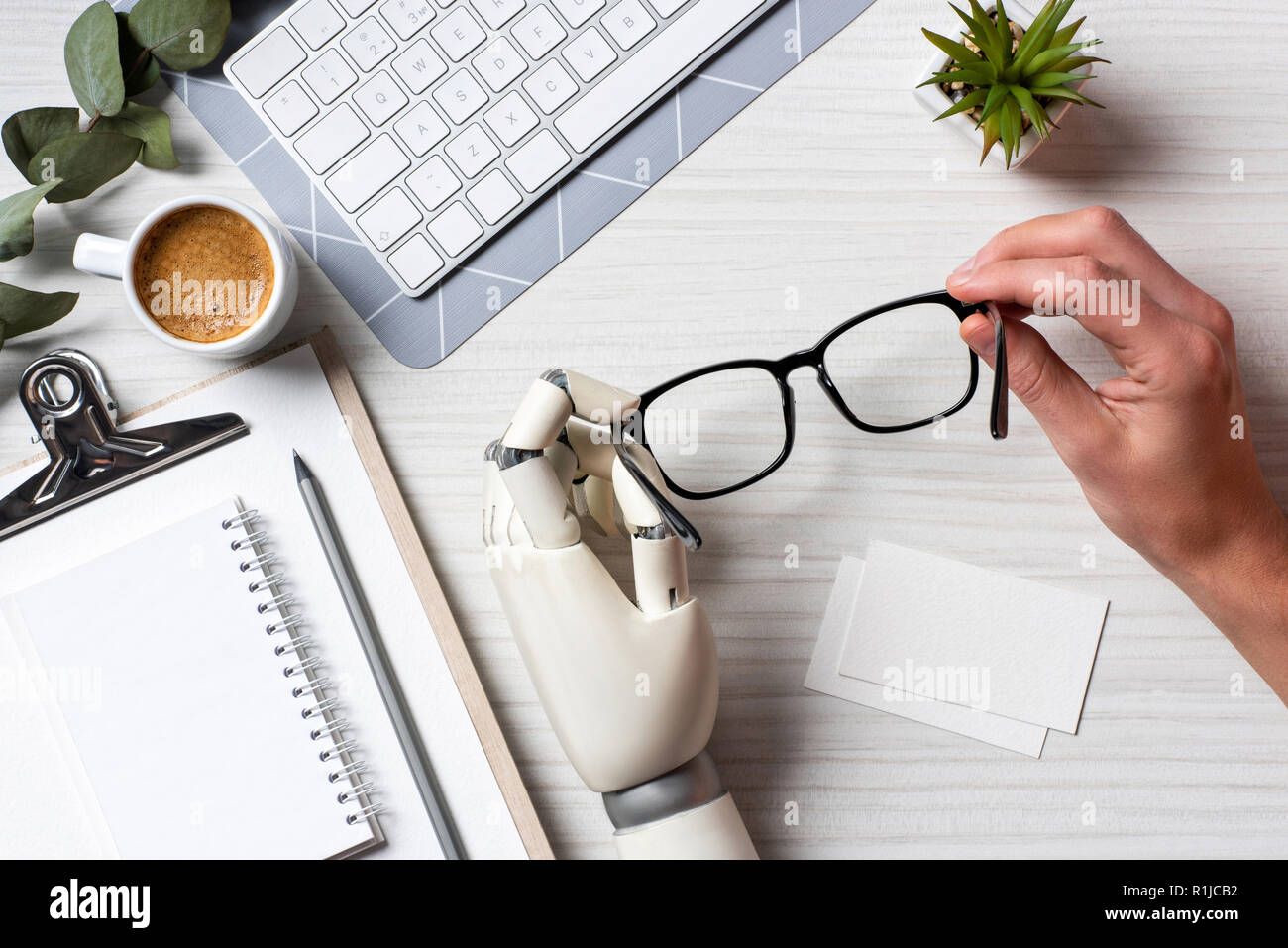 Immagine ritagliata di imprenditore con cyborg mano azienda occhiali a tavola con vuoto biglietti da visita e la tastiera del computer in ufficio Foto Stock