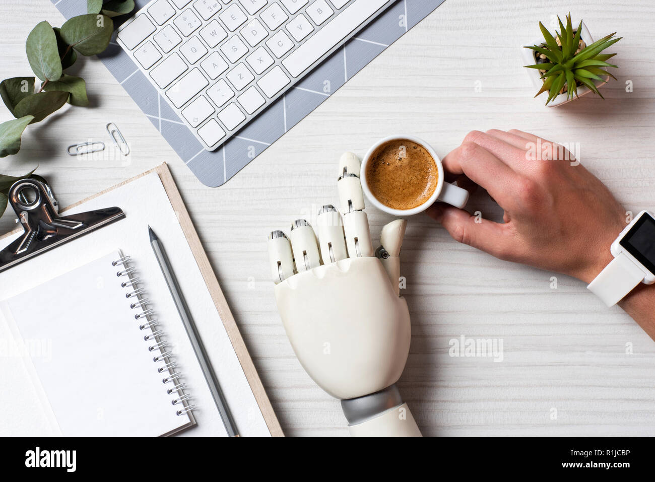 Immagine ritagliata di imprenditore con protesi mano e smartwatch seduta a tavola con la tazza di caffè e una tastiera di computer in ufficio Foto Stock