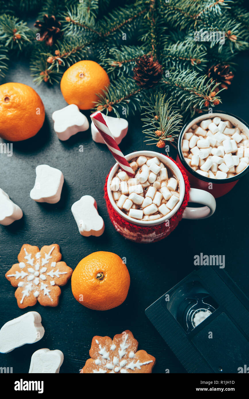 Vista dall'alto di tazze di bevande calde con marshmallows, cassette video e tangerini sulla superficie nera, natale colazione concept Foto Stock