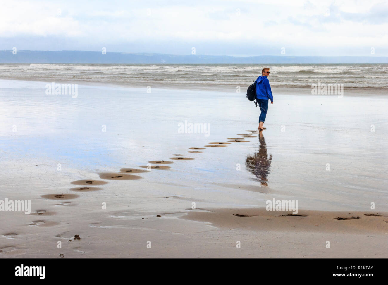 A piedi nudi donna in abiti casual camminare verso il mare sulla spiaggia spazzate dal vento a Westward Ho, Devon, Regno Unito Foto Stock