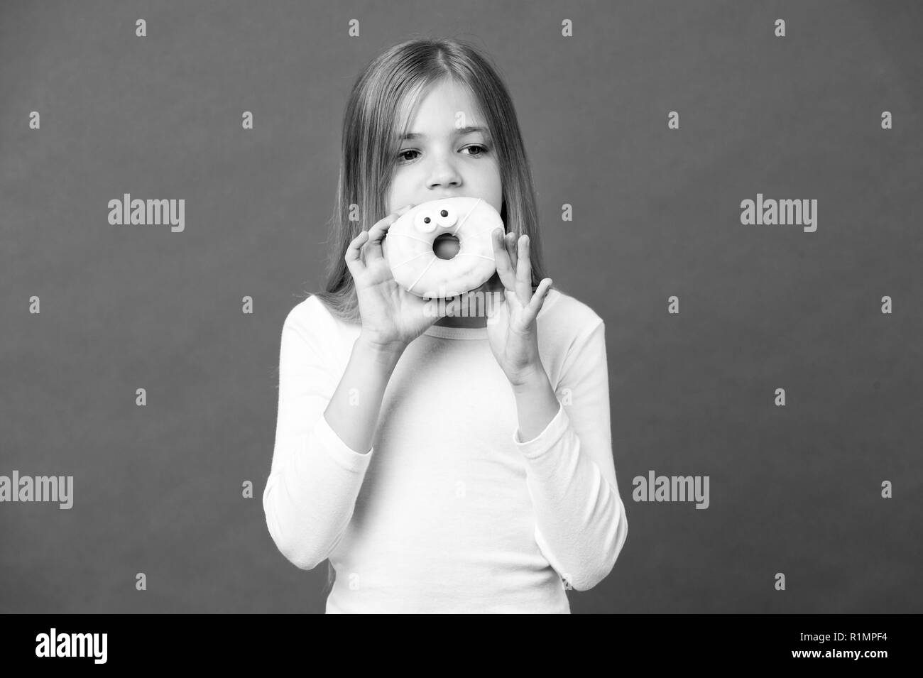 Ragazza con grande ciambella con glassa. Dolcetto, smaltate bagel con gli occhi. Kid eating funny dessert, infanzia dolce. Bambino con capelli lunghi che indossa il vestito bianco isolato su sfondo viola. Foto Stock